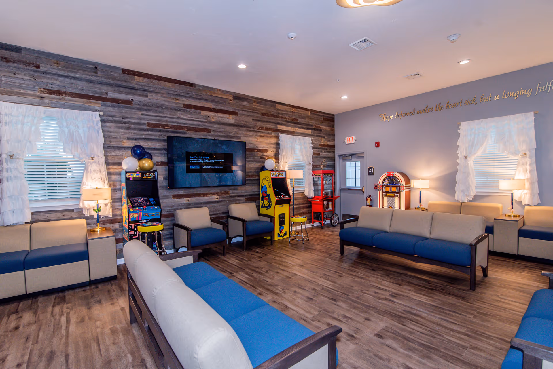 A spacious living room with multiple blue and beige cushioned sofas arranged around the room. The walls feature rustic wood paneling and light gray paint with white ruffled curtains on the windows. There is a flat-screen TV mounted on the wood-paneled wall, two vintage arcade game machines, a popcorn machine, and a jukebox. The room has wooden flooring and soft lighting from table lamps.