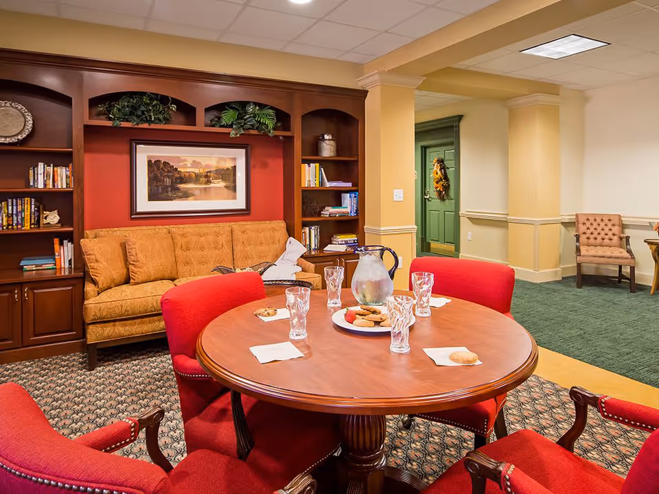 A cozy common area with a round wooden table surrounded by four red upholstered chairs. On the table are a pitcher of water, four glasses, a plate of cookies, and napkins. Behind the table is a tan patterned sofa set against a red accent wall with built-in wooden bookshelves filled with books and decorative items. A framed landscape painting hangs above the sofa. In the background, there is a green door with a wreath and a beige armchair against a cream-colored wall.