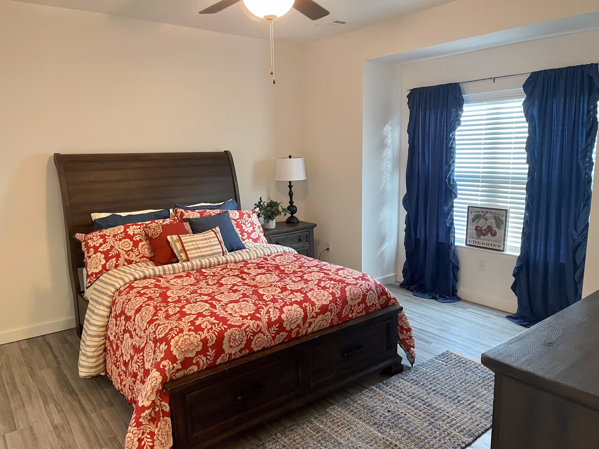 A bedroom with a wooden bed frame and a red floral patterned bedspread with multiple pillows. There is a nightstand with a lamp and a small plant next to the bed. A window with blue curtains and white blinds is on the right side, with a decorative sign on the windowsill. The floor is wood with a woven rug partially visible.