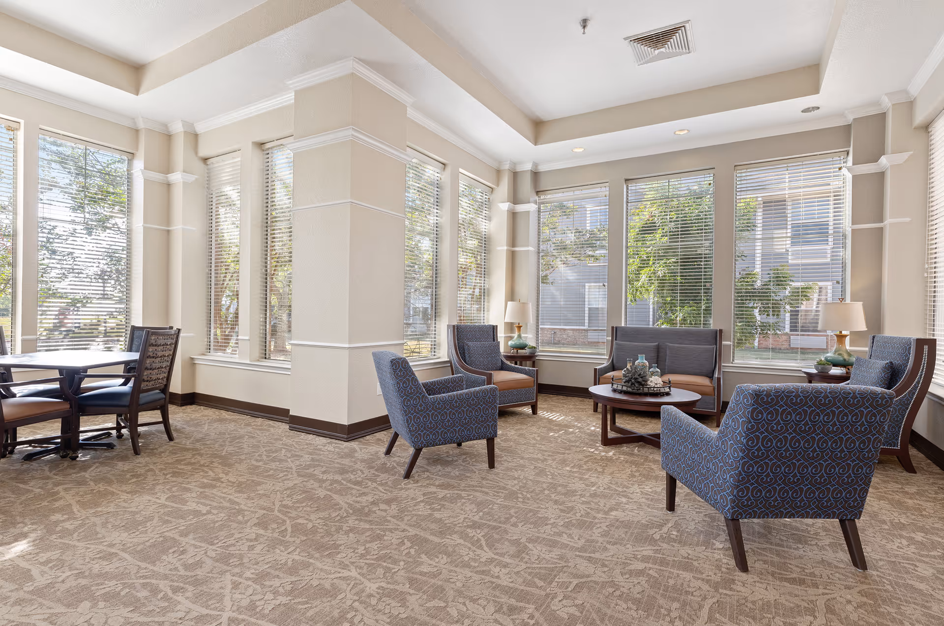 A bright and spacious living room area in a senior living facility with large windows letting in natural light. The room features several upholstered armchairs and a loveseat arranged around a round wooden coffee table with decorative items. There are two side tables with lamps, and a dining table with chairs is visible on the left side near the windows.