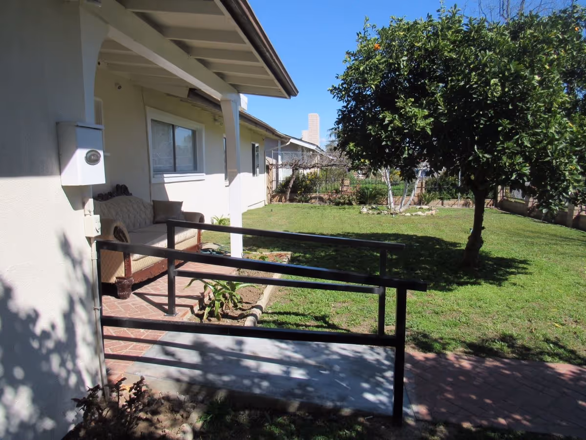 Outdoor view of a yard with a green lawn, a tree with some fruit, and a paved walkway leading to a small porch area with a cushioned bench. The porch has a black metal handrail and is attached to a light-colored building under a clear blue sky.