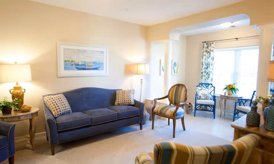 A bright and cozy living room with a blue couch, two patterned pillows, a wooden armchair with striped upholstery, two blue chairs near a window with floral curtains, a small round table with a plant, two lamps, and framed artwork of sailboats on the wall.