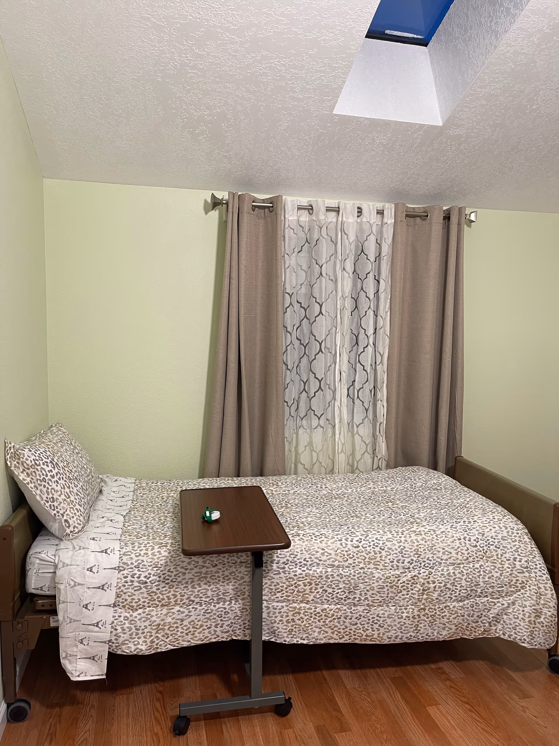 A single bed with leopard print bedding and a pillow is positioned against a light green wall. In front of the bed is a small overbed table with a few small items on it. Behind the bed, there is a window with sheer patterned curtains and beige drapes. The room has wooden flooring and a skylight in the ceiling.