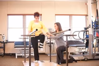 A woman in a yellow top and black pants is performing a physical therapy exercise by stepping on a platform while holding parallel bars. A physical therapist kneels beside her, providing support and guidance in a well-lit therapy room with exercise equipment and treatment tables in the background.