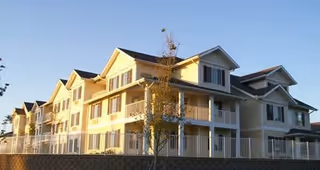 Exterior view of a multi-story residential building with balconies and a pitched roof, illuminated by sunlight in the late afternoon or early morning.