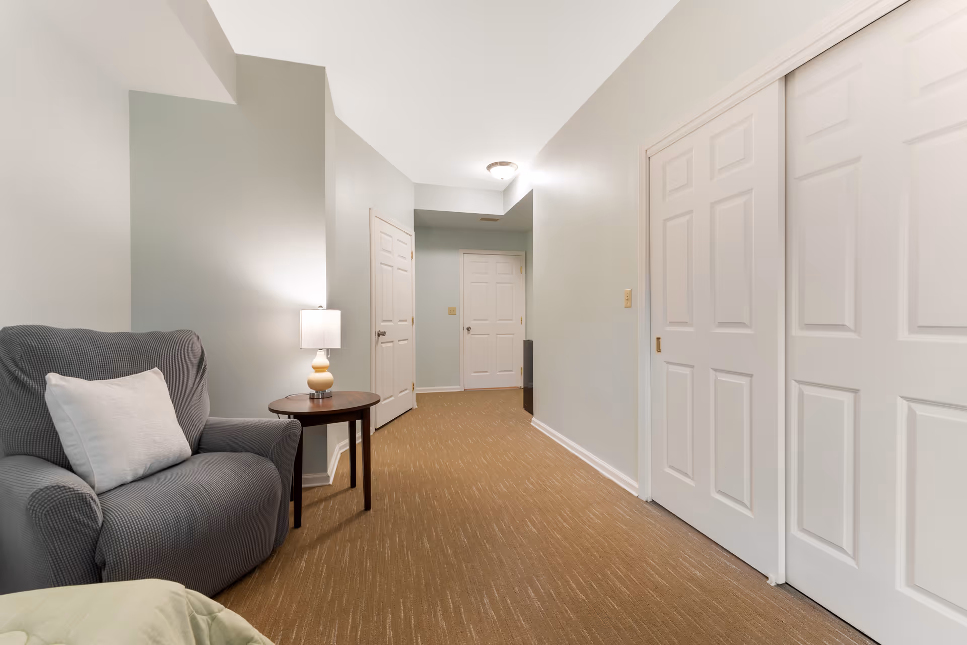 Interior hallway area with an upholstered armchair and side table lamp beside white closet and room doors on a beige carpet.