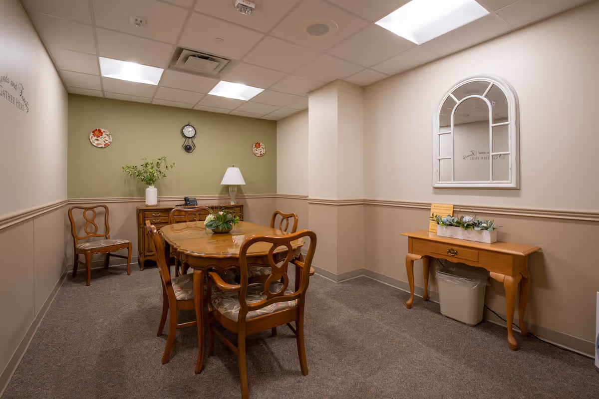 A small dining room with a wooden dining table surrounded by six chairs with cushioned seats. The walls are painted beige with a green accent wall at the far end. On the green wall, there is a clock and two decorative plates. A wooden sideboard with a lamp and a vase with greenery is placed against the green wall. On the right side, there is a wooden console table with a white planter containing green plants and a trash bin underneath. Above the console table is a decorative white-framed mirror. The room has a carpeted floor and a drop ceiling with fluorescent lights.