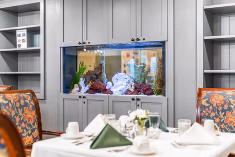 Dining area with a table set for a meal, featuring white tablecloth, folded green and white napkins, cups, glasses, and a small flower centerpiece. In the background, there is a built-in aquarium with colorful coral and plants, flanked by empty gray shelves on both sides.