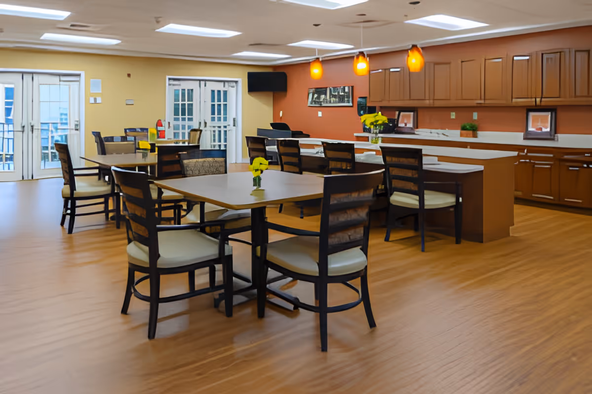 A spacious dining area with multiple tables and chairs arranged neatly. The room features a kitchen counter with bar stools, wooden cabinets, and pendant lights hanging from the ceiling. There are double glass doors leading outside, and small flower vases are placed on the tables.