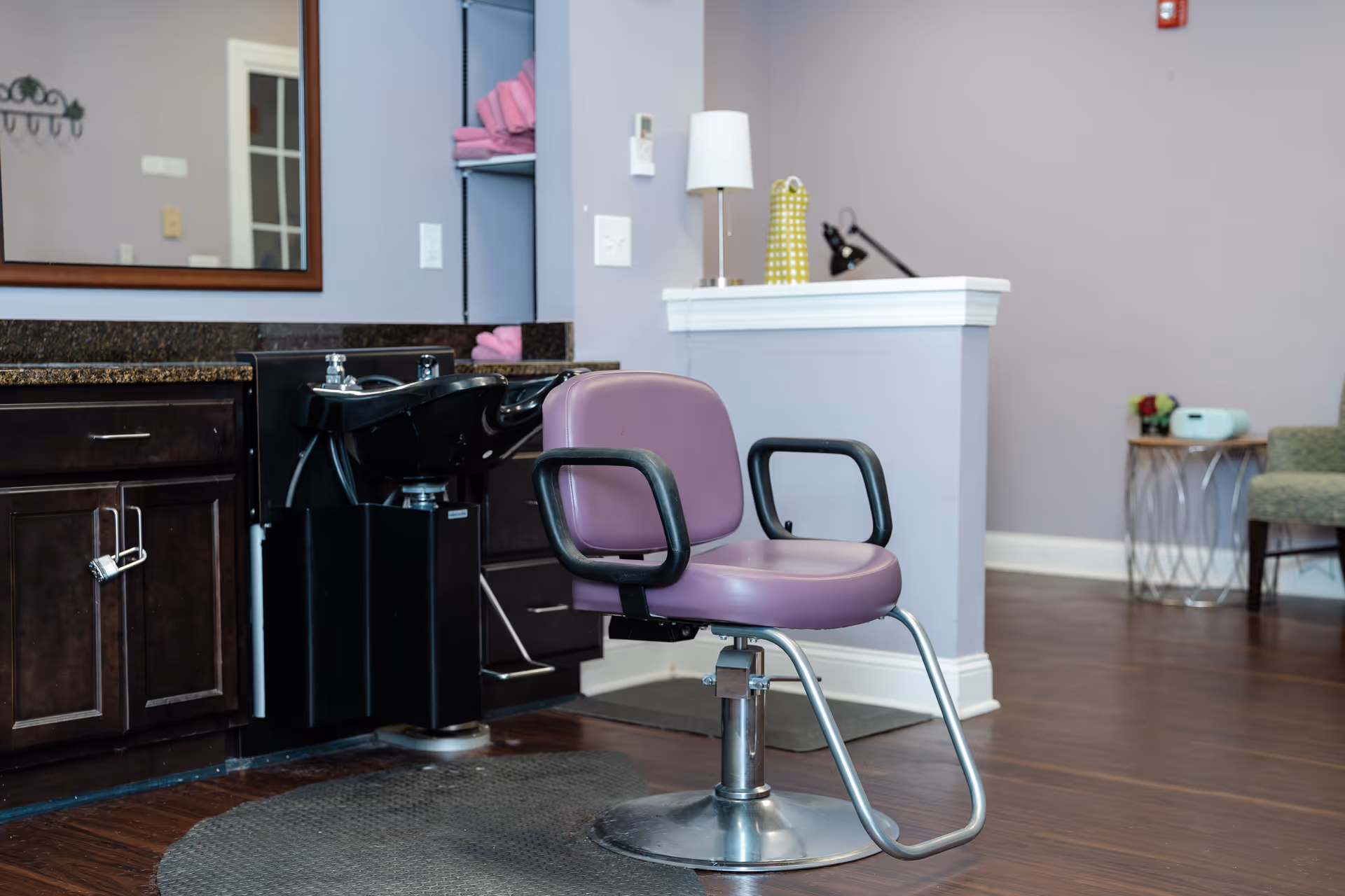 Interior view of a salon area in a senior living facility featuring a purple salon chair in front of a black hair washing station with cabinets underneath. A large mirror is mounted on the wall above the counter. In the background, there is a small table with a lamp and decorative items, and a green upholstered chair is partially visible.