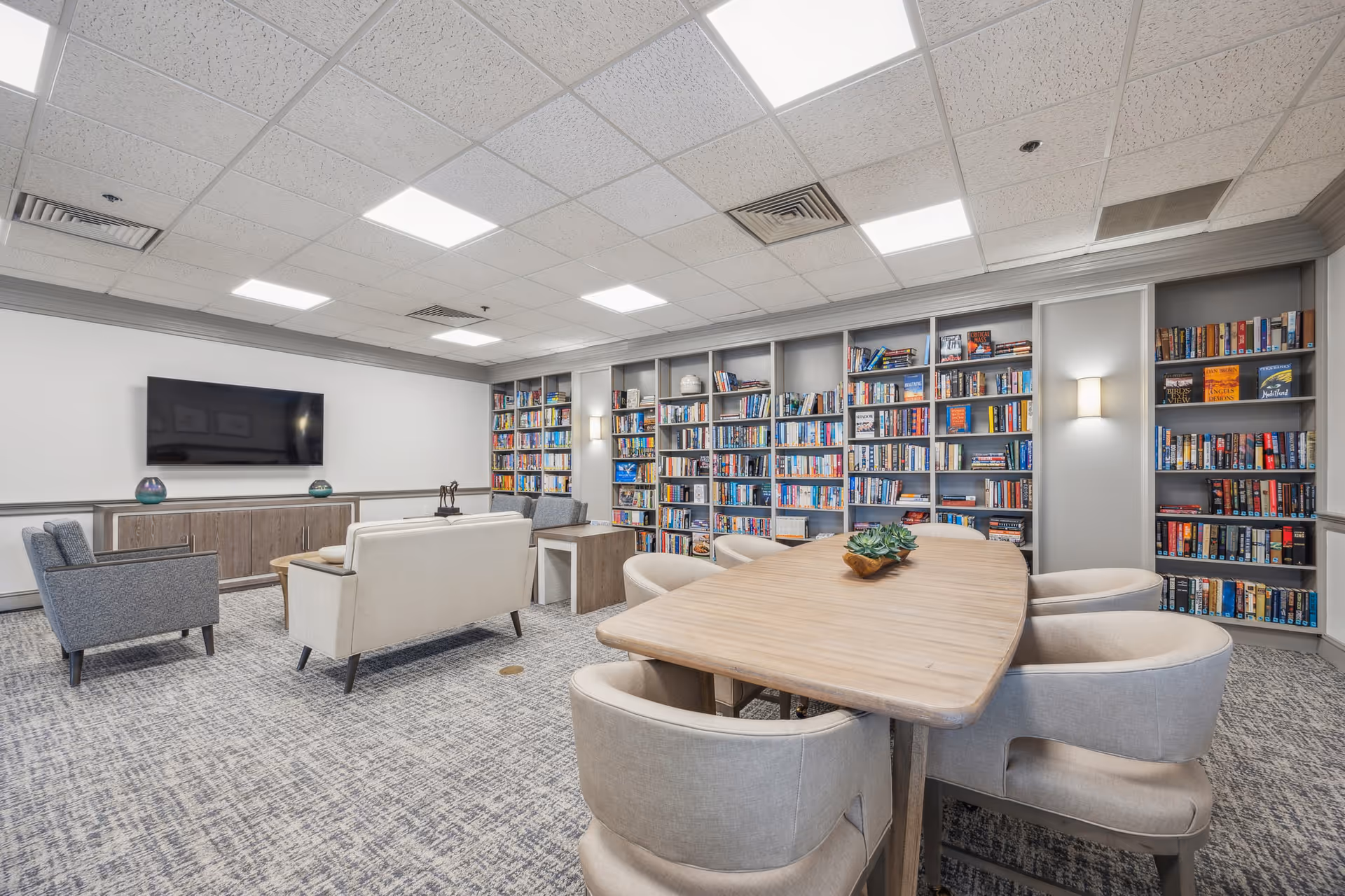 A bright and spacious library room with built-in bookshelves filled with books along the walls. The room features a large wooden table with six cushioned chairs around it, a seating area with a white sofa and two gray armchairs facing a wall-mounted flat-screen TV, and modern ceiling lights illuminating the space.