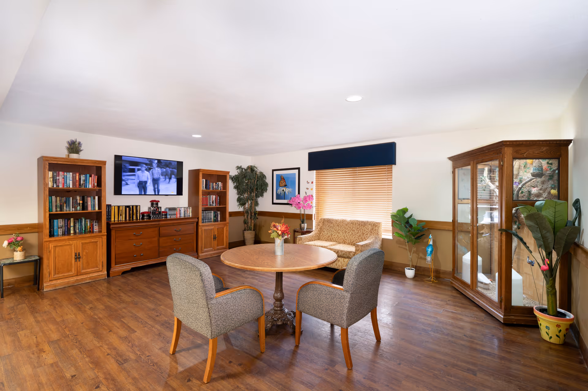 A cozy living room area with wooden flooring, featuring a round wooden table with two upholstered chairs around it. There is a small sofa against the wall under a window with wooden blinds and a dark blue valance. The room has wooden bookshelves filled with books and a flat-screen TV mounted on the wall. Various potted plants and flowers decorate the space, along with a wooden display cabinet on the right side.