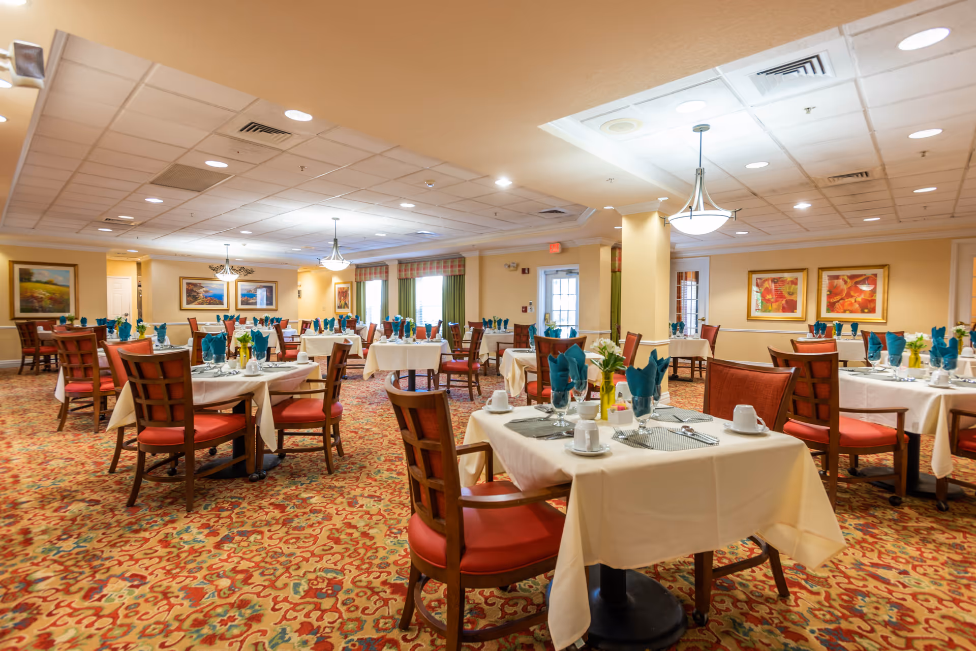 A spacious dining room with multiple tables covered in white tablecloths, each set with plates, cups, silverware, and teal folded napkins. The room has patterned carpet, warm yellow walls adorned with framed artwork, and large windows with green curtains allowing natural light. Ceiling lights and chandeliers provide additional illumination.