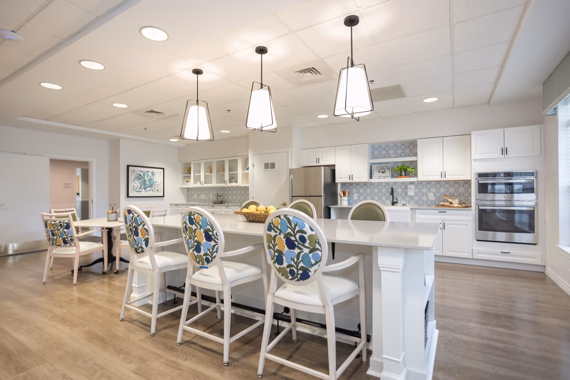 Bright and spacious kitchen area in a senior living facility with a large white island countertop surrounded by chairs with floral patterned backs. The kitchen features white cabinets, stainless steel refrigerator, double oven, and blue patterned backsplash tiles. Three pendant lights hang above the island, and there is a small table with chairs in the background.