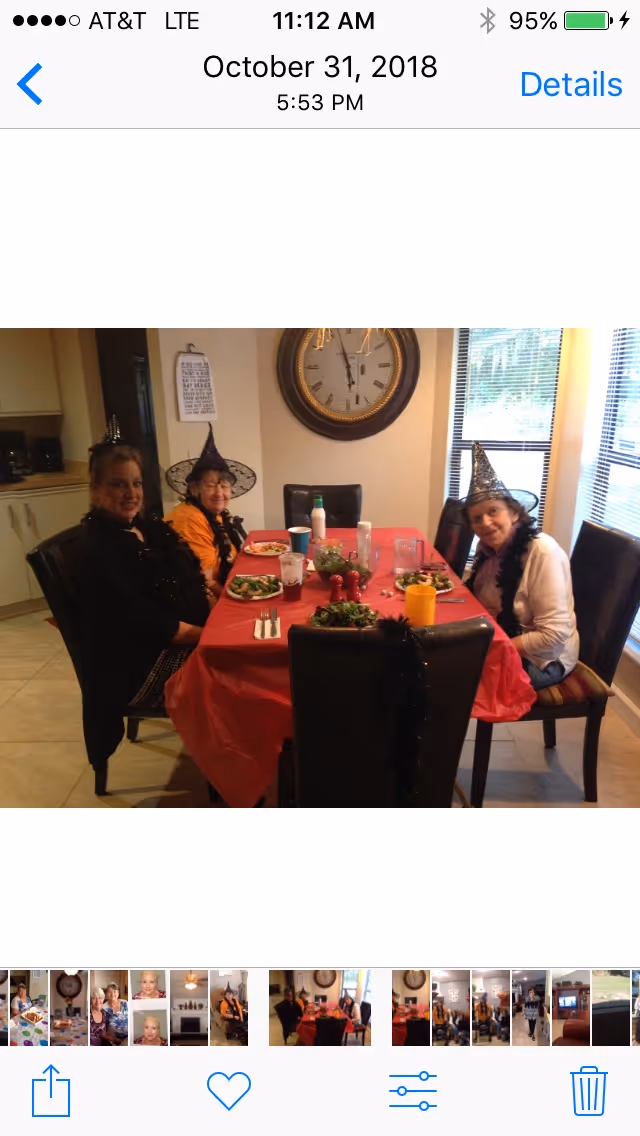 Three women wearing witch hats sitting around a dining table covered with a red tablecloth, with plates of food and drinks in front of them. The room has a large wall clock and windows with blinds.