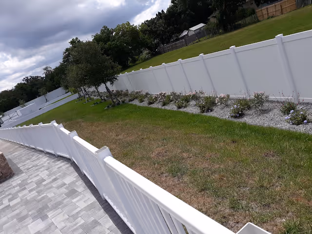 Sloped backyard with a white vinyl fence, paved walkway, grassy lawn and planted beds under a cloudy sky.