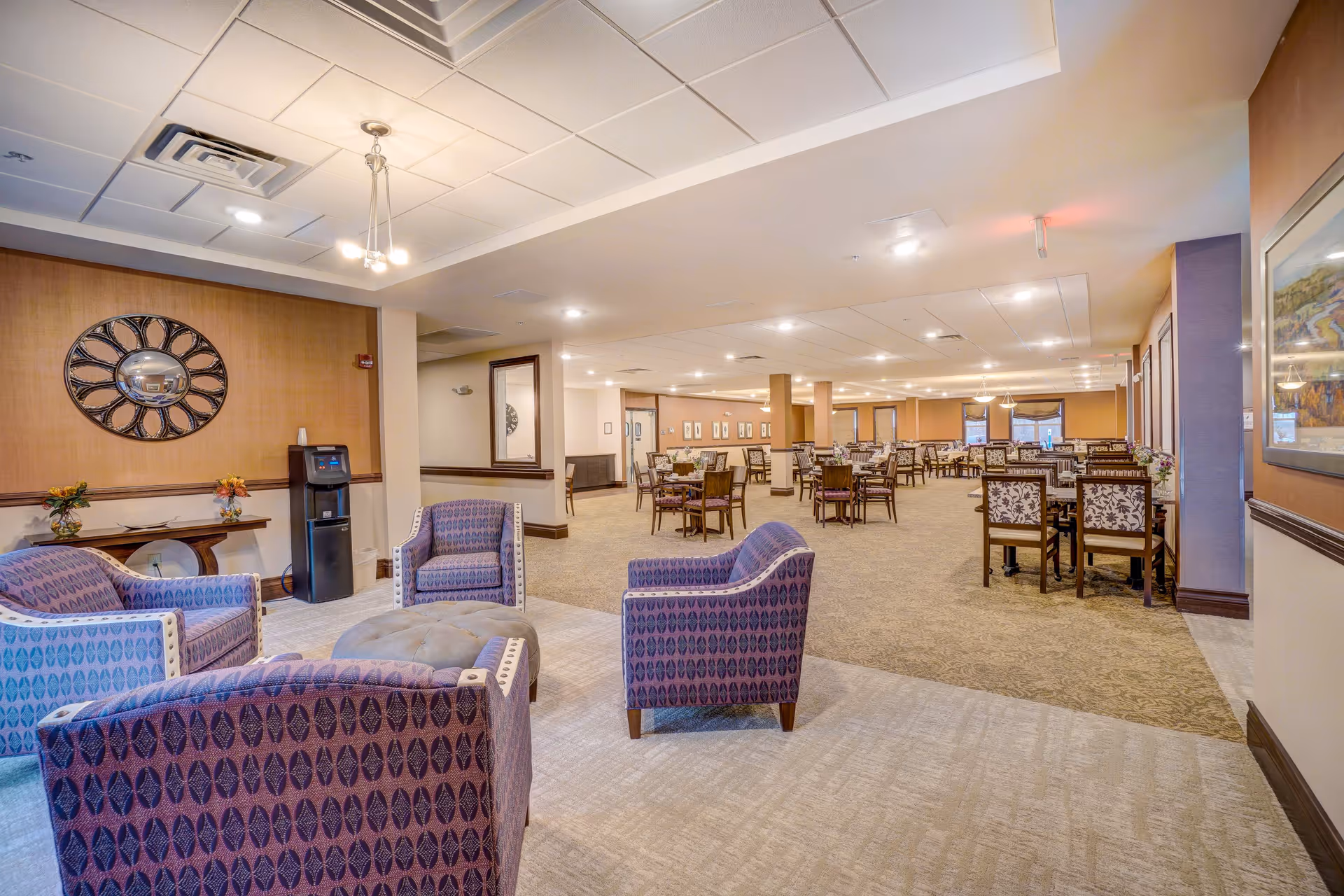 A spacious senior living facility common area with a seating arrangement of four purple patterned armchairs around a round ottoman in the foreground. In the background, there are multiple dining tables and chairs arranged neatly across the room. The walls are decorated with framed artwork and a large decorative mirror. The room is well-lit with ceiling lights and has a warm, inviting atmosphere.