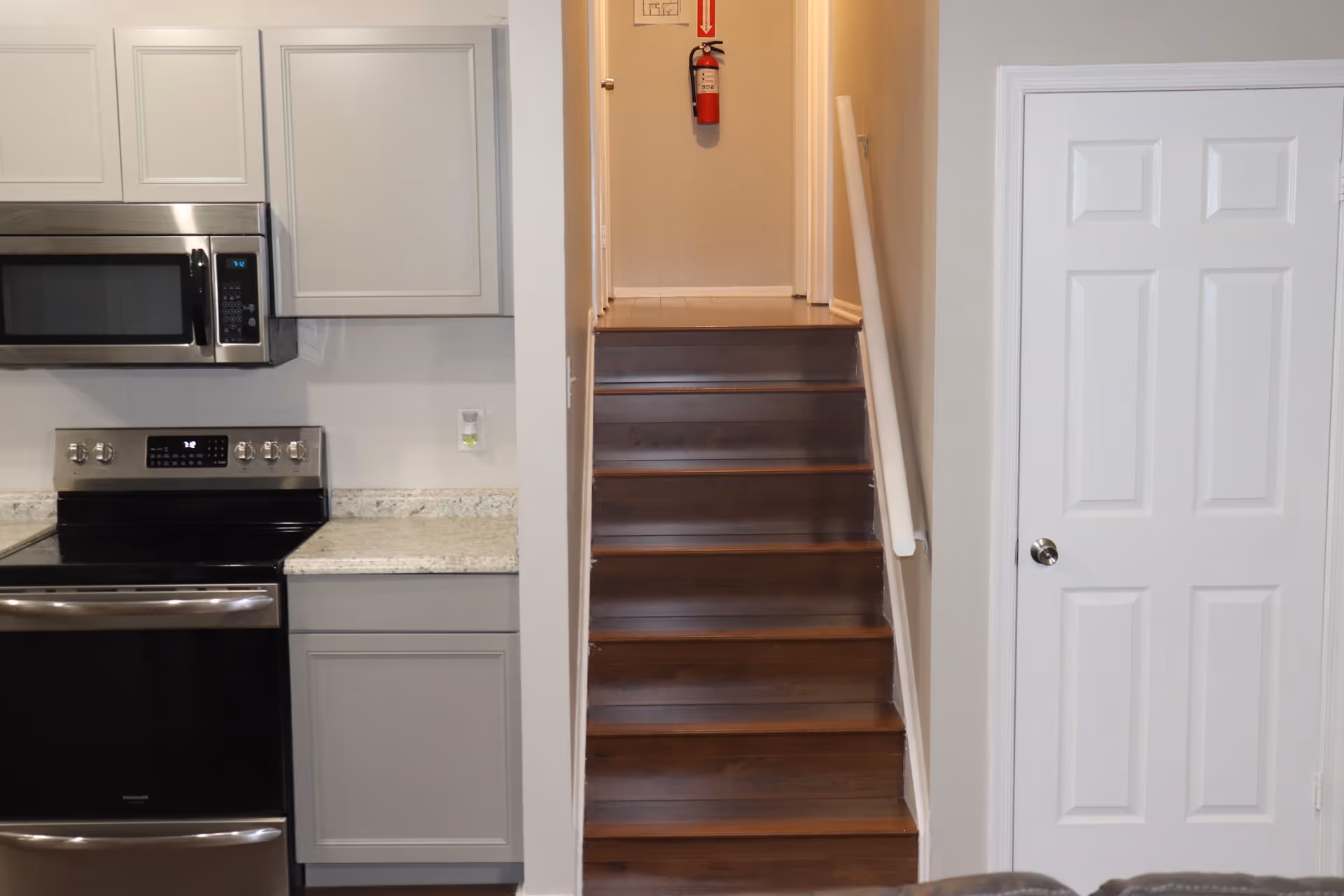 Interior view of a home showing a kitchen area with a stainless steel microwave and stove on the left, a staircase with wooden steps and a white handrail in the center, and a white door on the right.