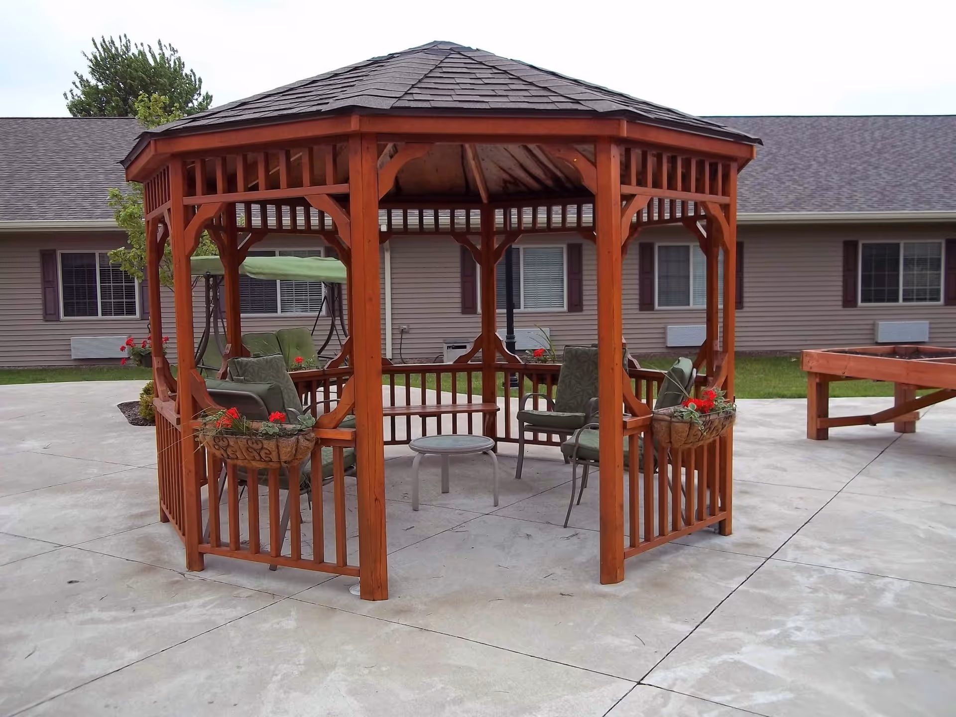 Wooden gazebo with chairs and potted flowers in a paved courtyard outside a senior living building.
