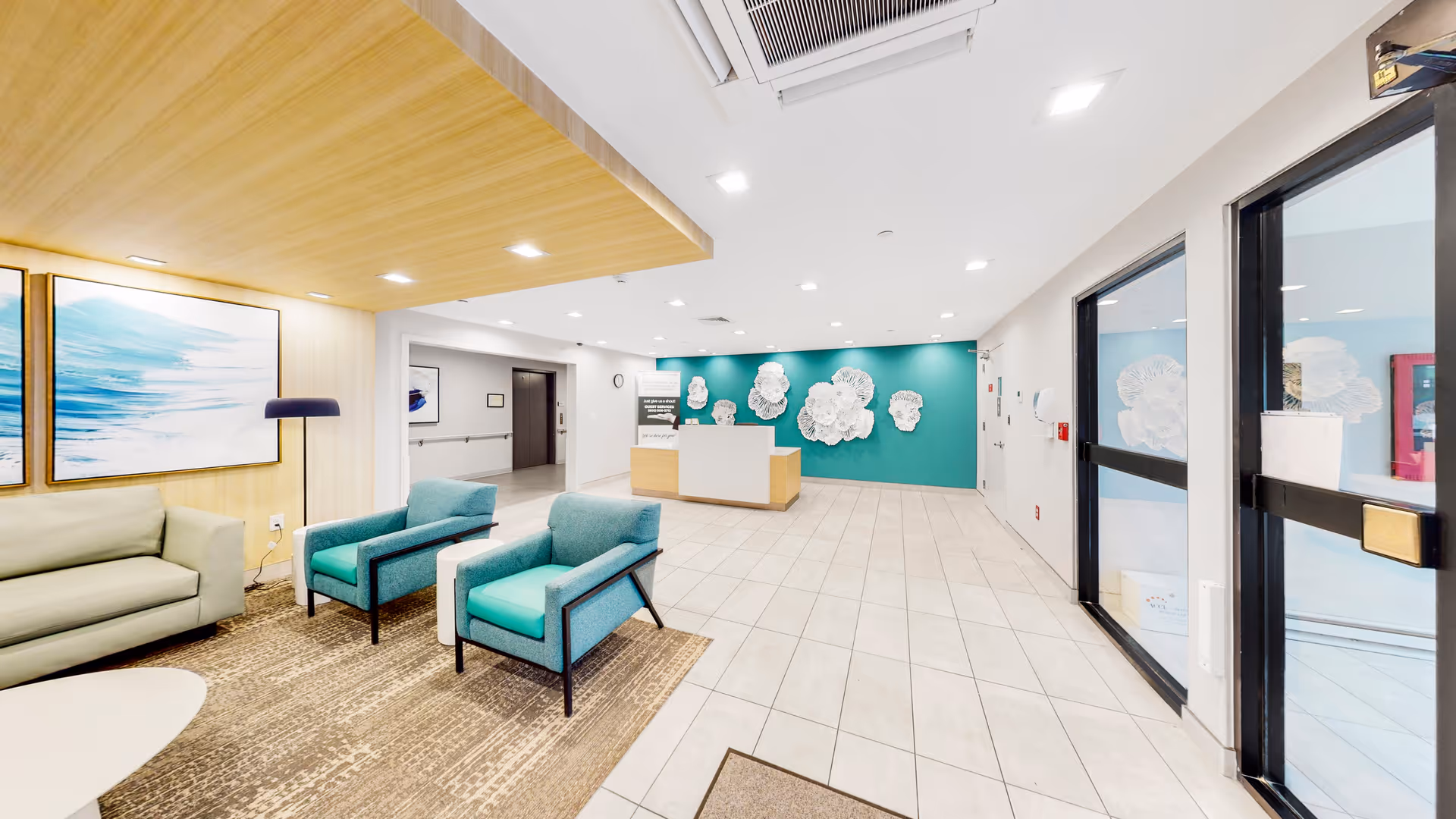 A bright and modern reception area with a white reception desk against a teal accent wall decorated with white floral wall art. The room features light-colored tiled flooring, a seating area with a green sofa and two teal armchairs on a brown rug, and large glass doors on the right side. The ceiling has recessed lighting and a wooden panel section above the seating area.