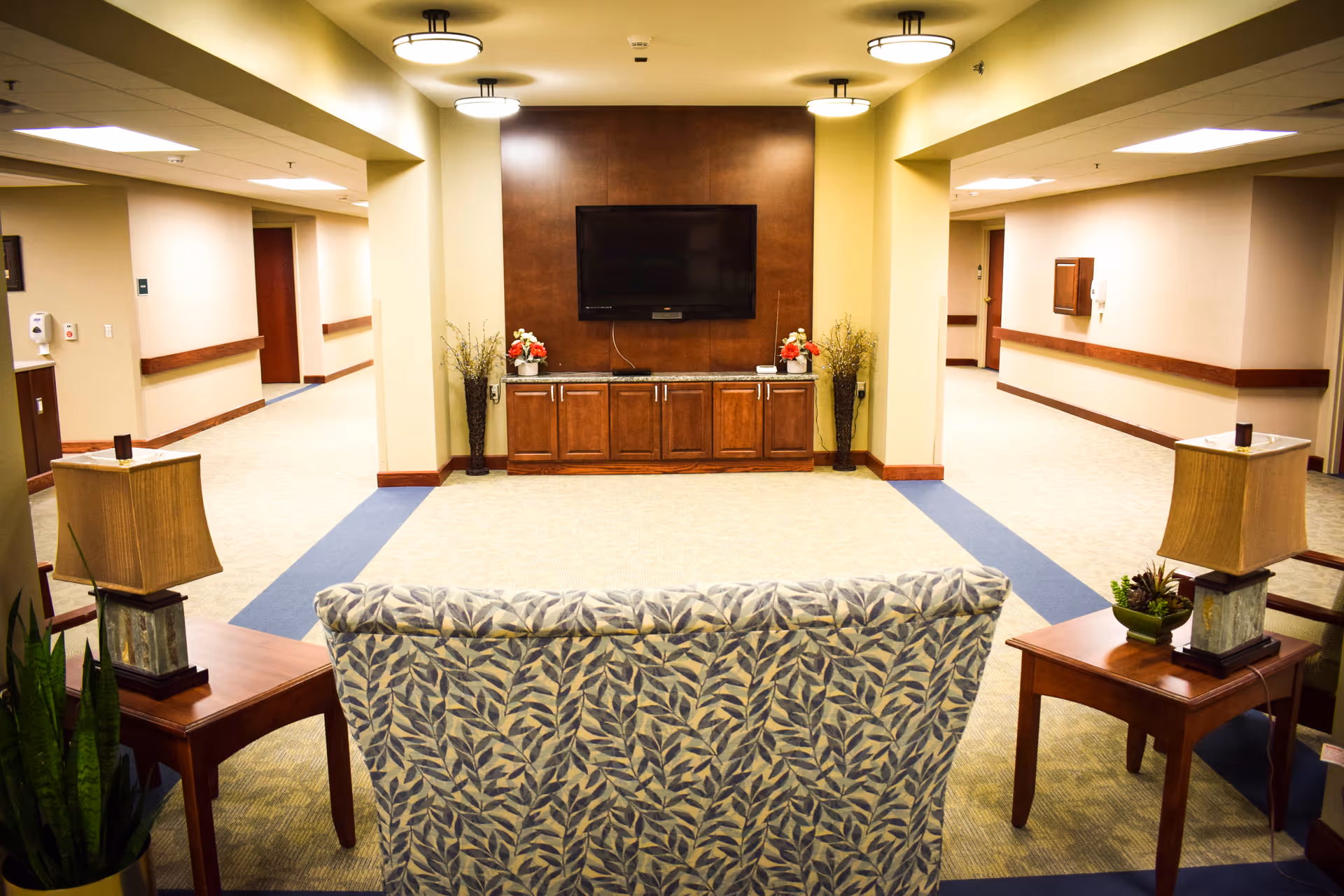 Seating area in a senior living facility with patterned chairs facing a wall-mounted TV and wooden console, flanked by side tables and lamps.