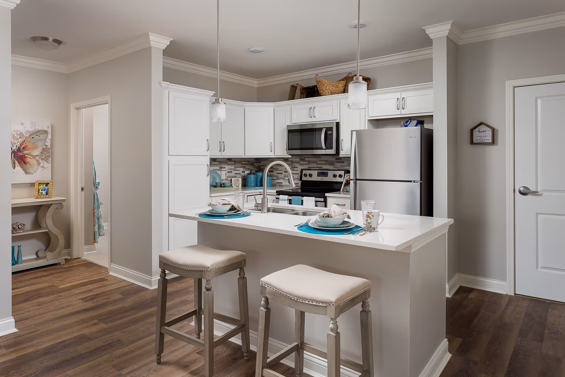 A modern kitchen with white cabinetry, stainless steel appliances including a refrigerator, microwave, and stove. There is a white island with a sink and two cushioned bar stools. The kitchen has pendant lights hanging above the island and a wooden floor. A small table with decorative items and a butterfly painting is visible in the adjacent hallway.