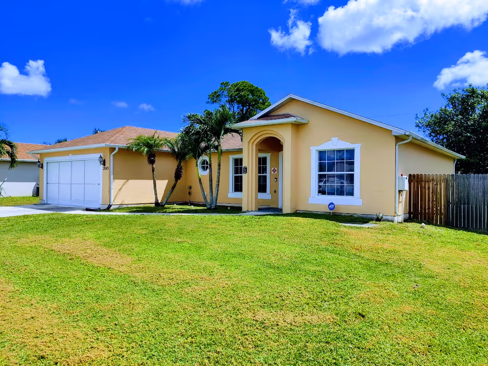 Single-story yellow house with an attached garage, front lawn and palm trees under a bright blue sky.