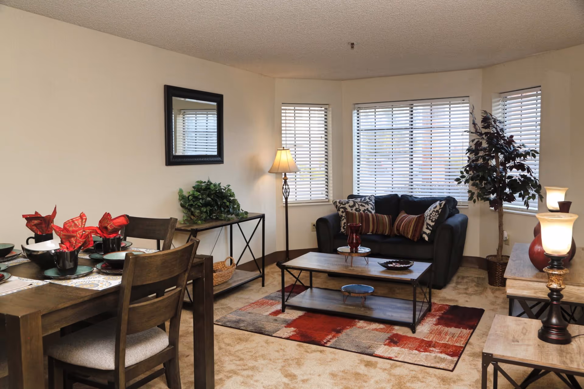 A cozy living and dining area with a dark wooden dining table set with black dishes and red napkins, a black couch with patterned cushions, a wooden coffee table on a red and beige rug, a floor lamp, a potted plant, and a side table with a lamp. The room has beige walls and carpet, and windows with white blinds.