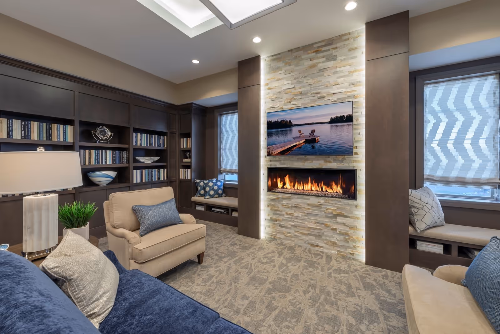 A cozy living room with a beige armchair and a blue sofa adorned with pillows. The room features a modern fireplace set in a stone wall with a flat-screen TV mounted above it displaying a serene lake scene. Built-in dark wood bookshelves filled with books and decorative items line one wall, and two windows with patterned blinds flank the fireplace. The room is softly lit with recessed ceiling lights and a table lamp.