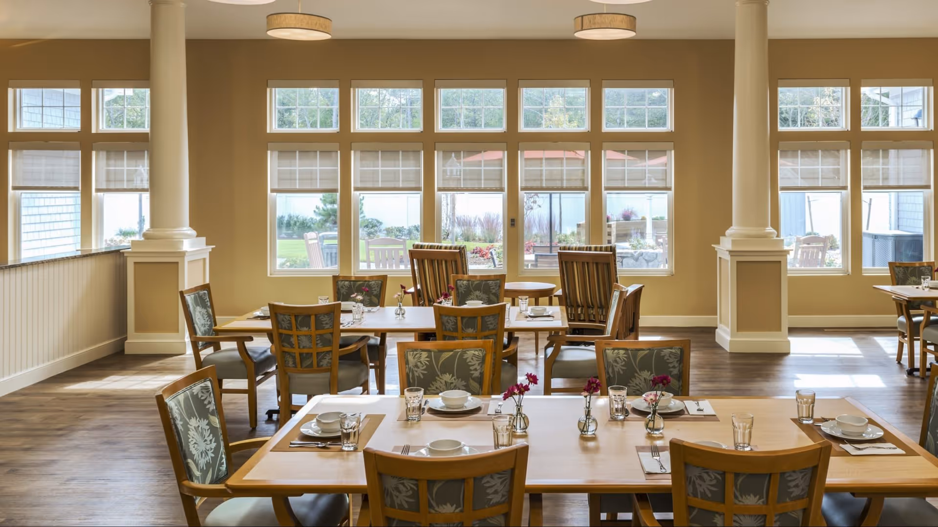 A bright dining room with wooden tables and chairs arranged neatly. Each table is set with white plates, bowls, glasses, and silverware, along with small vases holding purple flowers. Large windows line the back wall, allowing natural light to fill the room and providing a view of an outdoor garden area.
