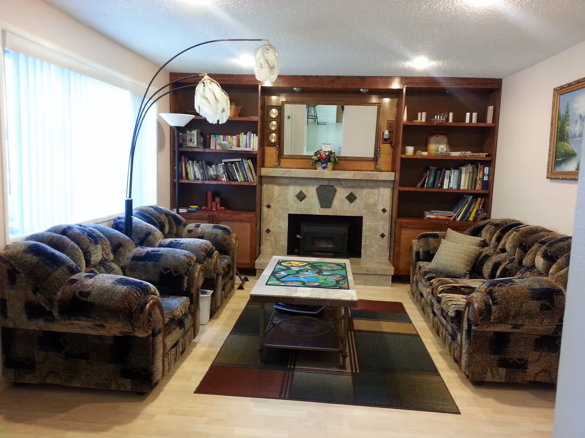 A cozy living room with two patterned sofas facing each other across a coffee table with a colorful board game on it. Behind the coffee table is a fireplace with a tiled surround and a large mirror above it. On either side of the fireplace are wooden bookshelves filled with books and decorative items. A tall floor lamp with three curved arms and white lampshades stands next to one sofa. A large window with vertical blinds lets in natural light from the left side.