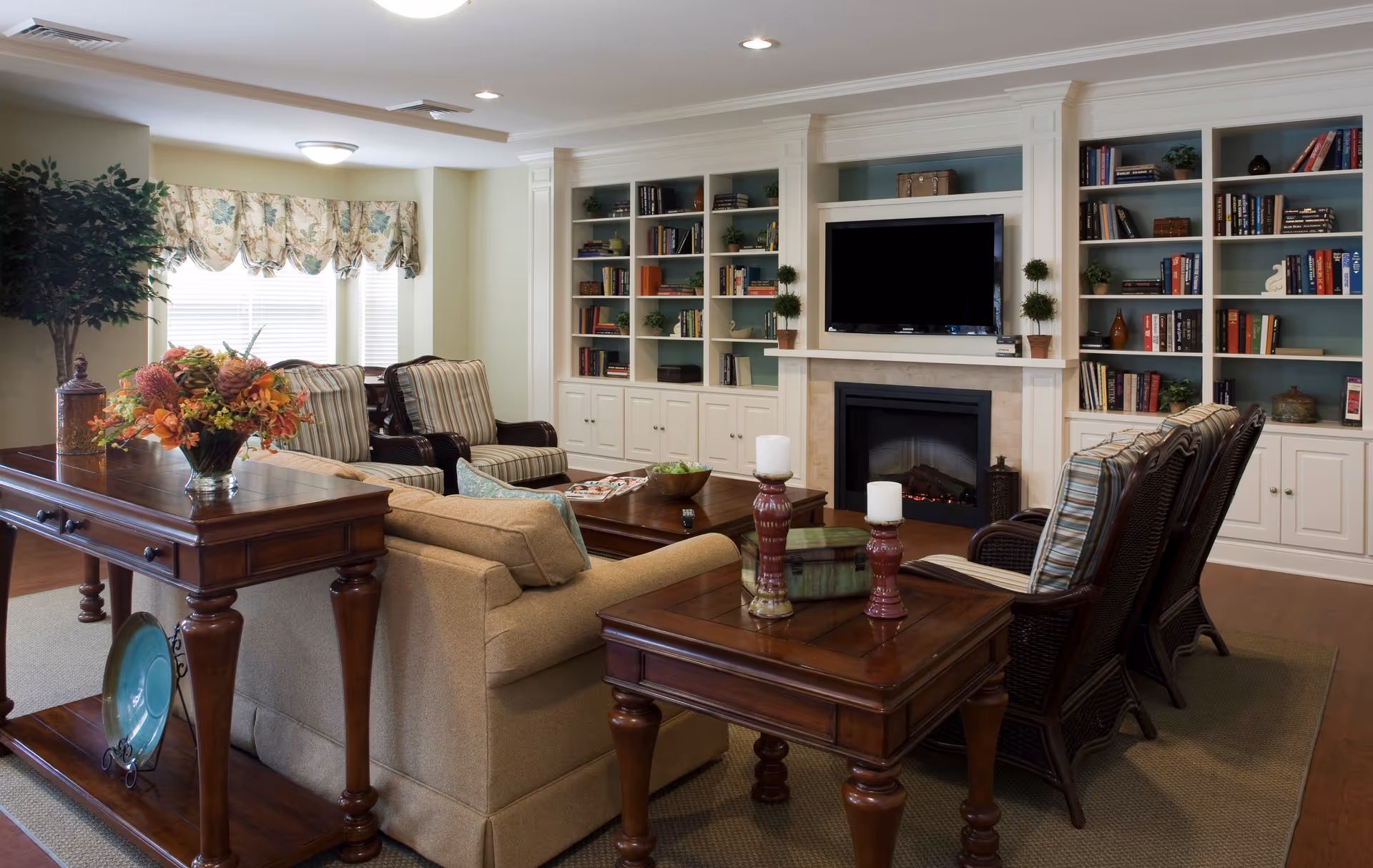 Cozy furnished living room with sofas and armchairs arranged around wooden tables, built-in bookshelves, a fireplace, and a wall-mounted TV.