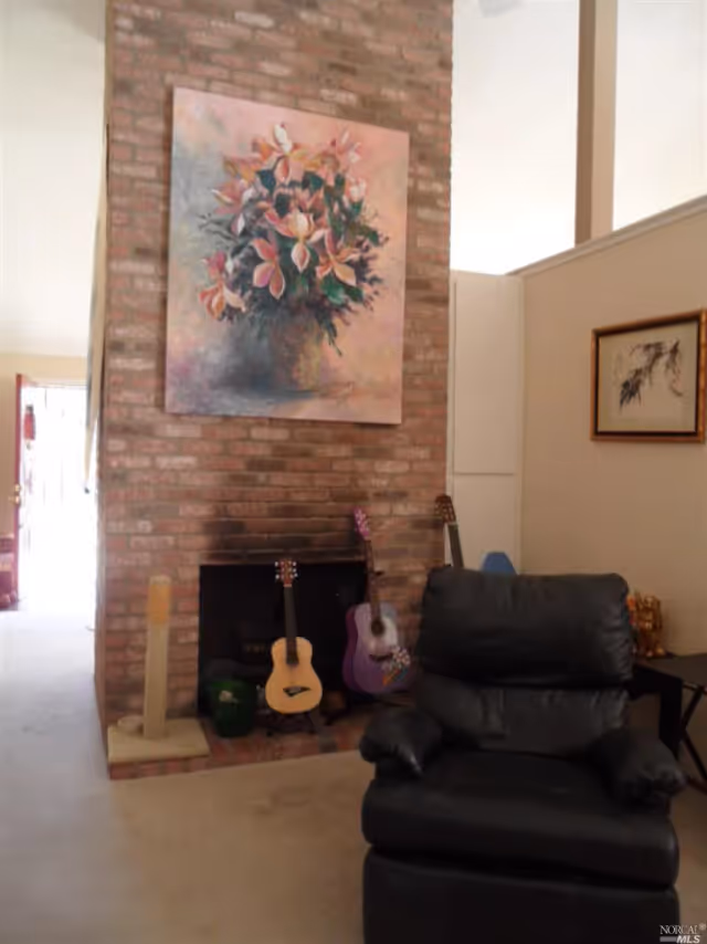 Interior view of a living room featuring a brick fireplace with three guitars placed inside it. Above the fireplace hangs a large painting of a floral bouquet. In front of the fireplace is a black leather recliner chair. To the right, there is a small table with a framed artwork hanging on the wall above it. The room has light-colored walls and carpeted floor.