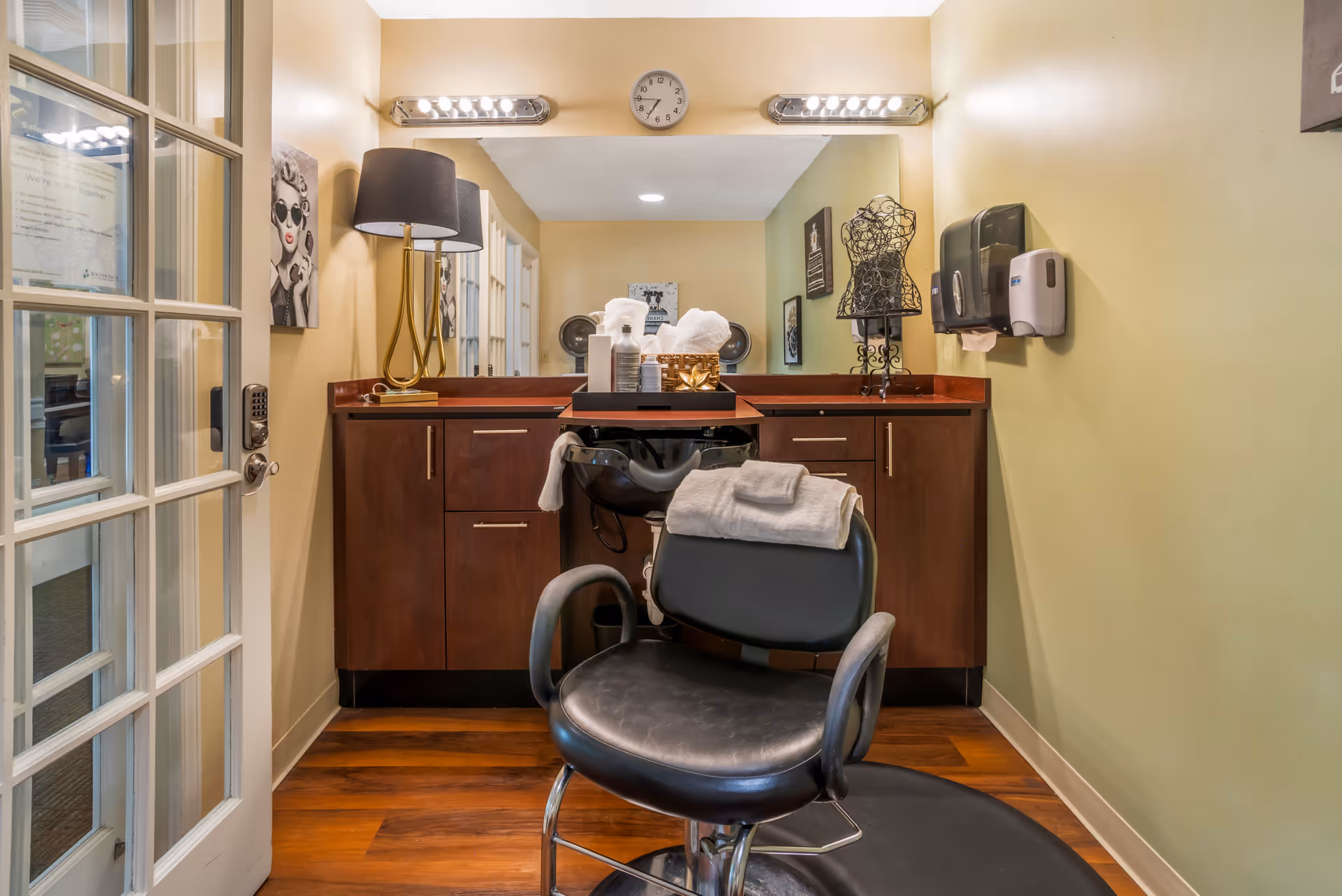 Interior view of a salon area in a senior living facility with a black salon chair in front of a wooden counter and mirror. The counter has various hair care products, towels, and a decorative lamp. The walls are painted light yellow and green, and there are framed pictures and a clock above the mirror. A glass door is visible on the left side.