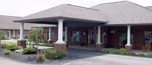 Exterior view of a single-story brick building with a covered entrance supported by white columns, surrounded by landscaping with shrubs and small trees.