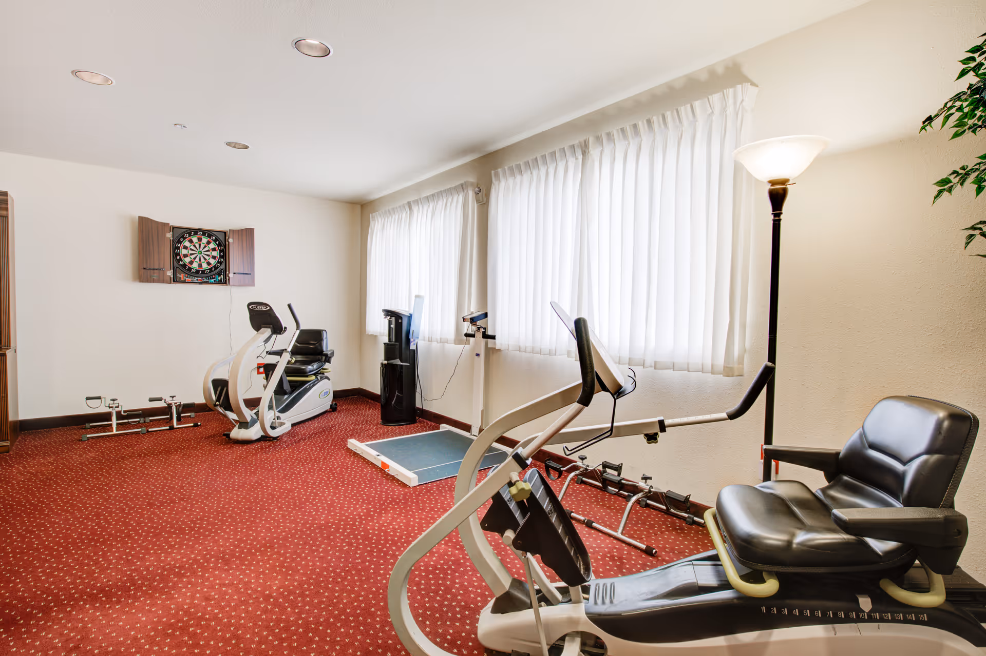 A bright indoor exercise room with recumbent bikes, small weights, a dartboard on the wall, and large curtained windows.