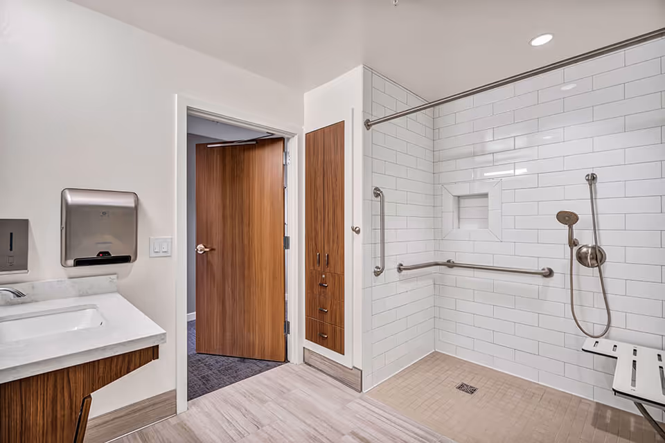 A modern bathroom featuring a walk-in shower with white subway tiles, grab bars, a handheld showerhead, and a fold-down shower seat. There is a wooden cabinet and a sink with a marble countertop on the left side. The bathroom door is open, revealing a carpeted room outside.