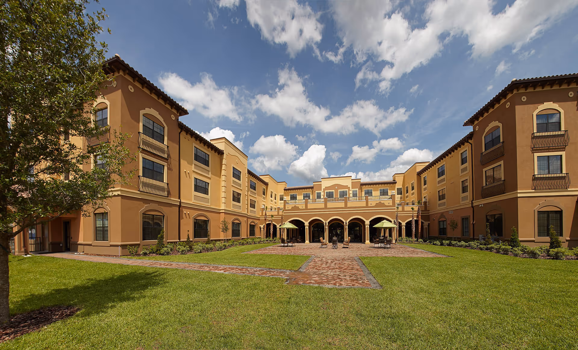 Exterior view of a large, three-story senior living facility building with a courtyard featuring green grass, brick pathways, outdoor seating with umbrellas, and a partly cloudy sky overhead.