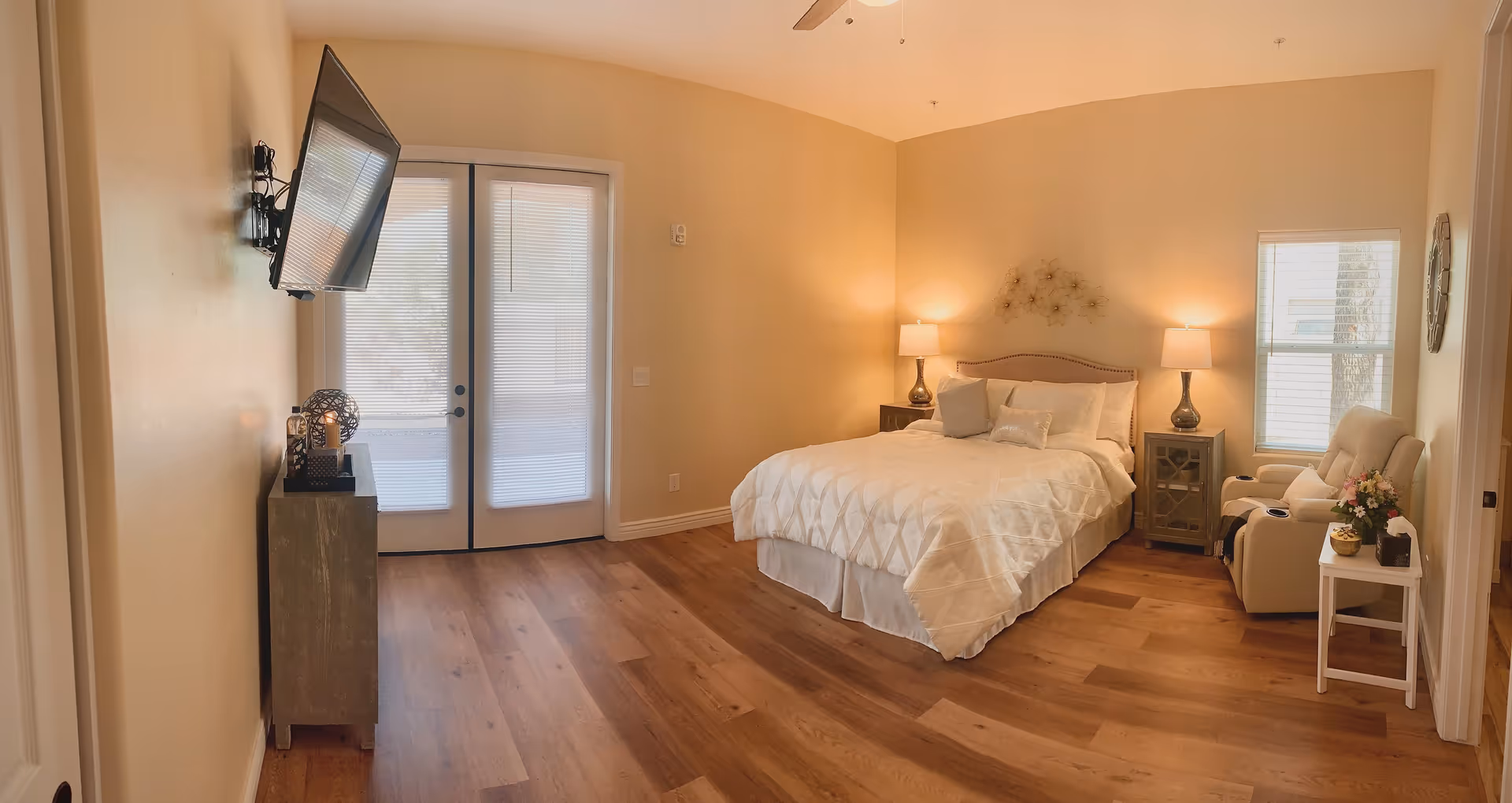A cozy bedroom with a large bed covered in white bedding, two bedside tables each with a lamp, a wall-mounted TV, a small cabinet, a reclining chair with a side table holding flowers, and double glass doors with blinds leading outside.