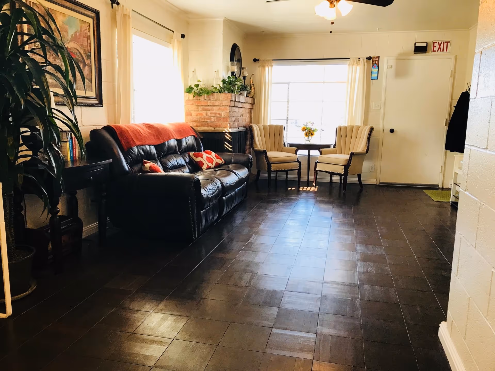 A cozy living room with a dark leather sofa adorned with red and patterned cushions, a red throw blanket, two beige armchairs around a small table with a flower vase, a brick fireplace with plants on top, a large window with white curtains, and a door with an exit sign above it. There is a large potted plant and a framed painting on the wall.