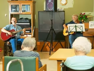 Two elderly men playing acoustic guitars and singing in a common room with an audience of elderly people seated and watching them. The room has a TV on a stand, a wooden cabinet, and a clock on the wall.