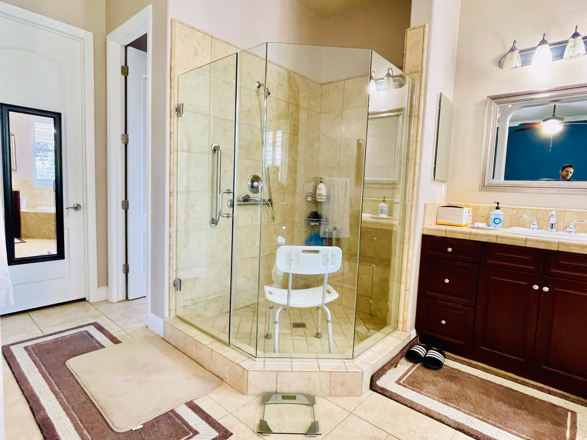 A bathroom with a glass-enclosed corner shower featuring beige tiles and a white shower chair inside. To the right is a dark wood vanity with a large mirror above it and two light fixtures. There are two brown and beige rugs on the tiled floor, a pair of black and white slippers, and a scale in front of the shower. A door with a full-length mirror is visible on the left side of the image.