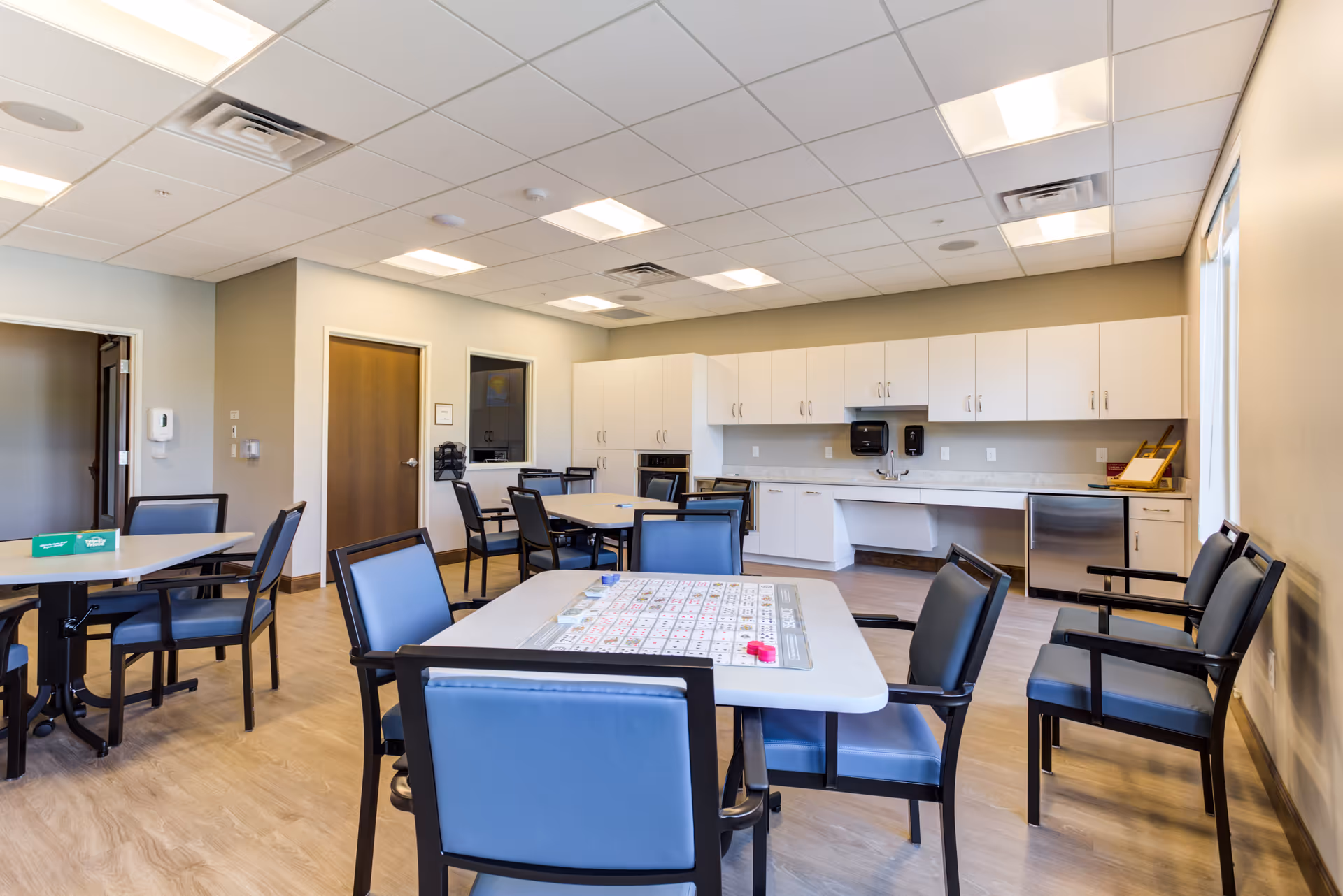 A bright room with multiple tables and blue cushioned chairs arranged around them. One table in the foreground has a large game board on it. The room has a light wood floor, white cabinets along one wall, a sink, and a small refrigerator. The ceiling has recessed lighting and a vent. There is a door and a window on one wall.