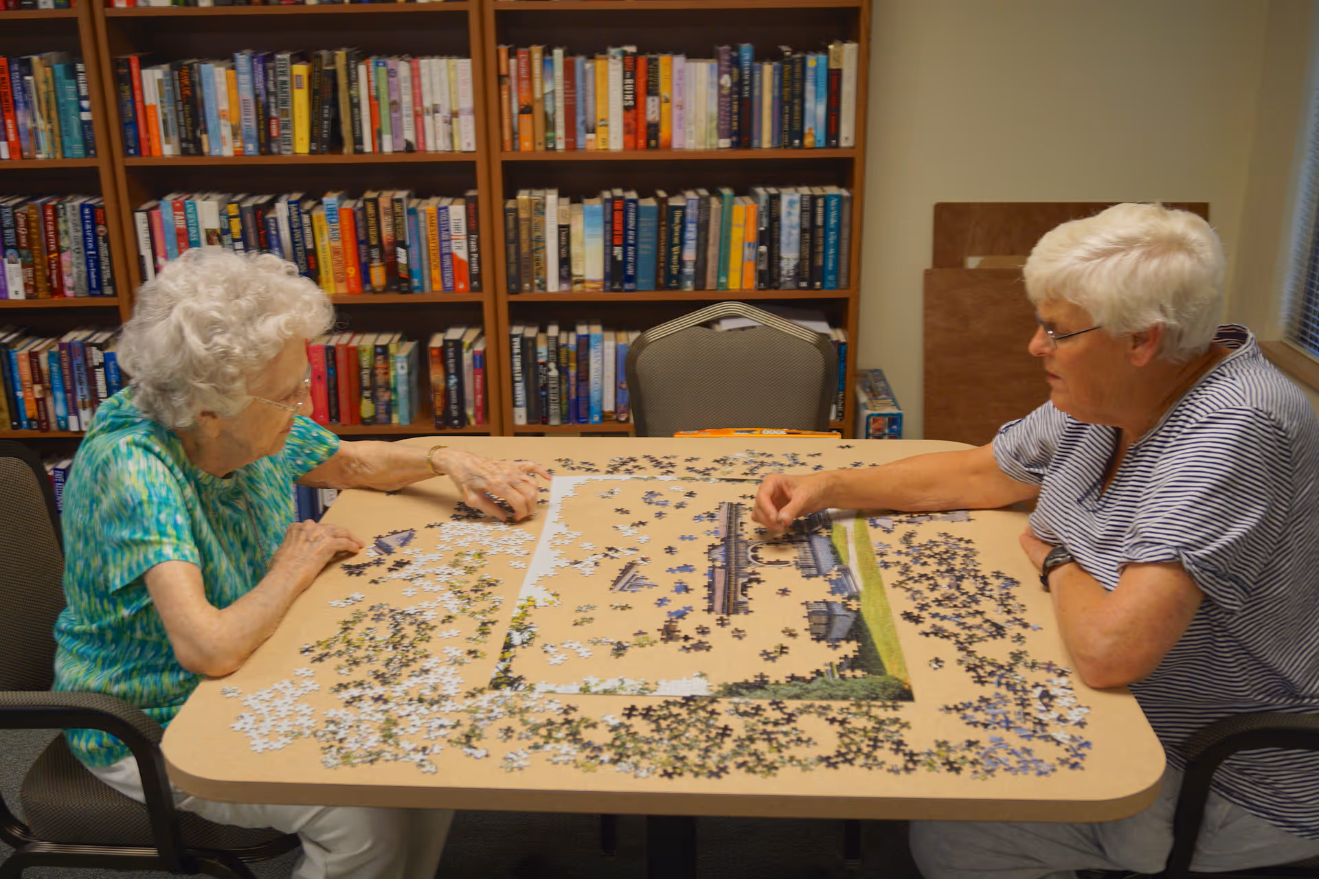 Two elderly women sitting at a table working on a large jigsaw puzzle in a room with bookshelves filled with books in the background.