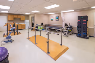 A rehabilitation therapy room with parallel bars for walking exercises, exercise equipment including a stationary bike and a purple exercise ball, a treatment table, and a kitchenette area with cabinets and a refrigerator. The room has bright overhead lighting and light-colored walls and flooring.