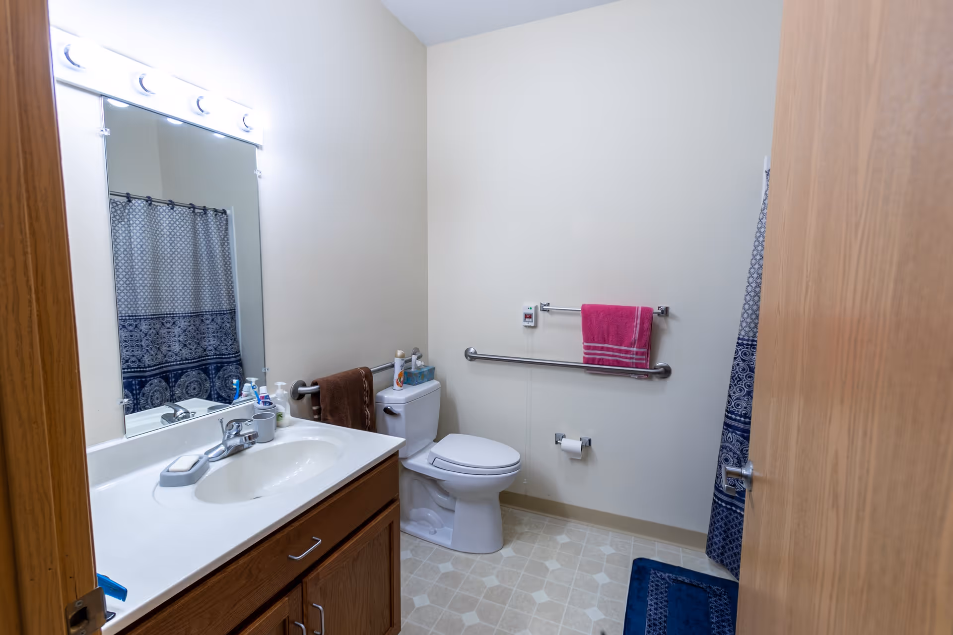 A bathroom with a white sink and countertop, wooden cabinet below, a large mirror with lights above it, a toilet with a brown towel hanging on the side, a pink towel hanging on a grab bar on the wall, a toilet paper holder, and a shower with a blue patterned curtain and matching bath mat.