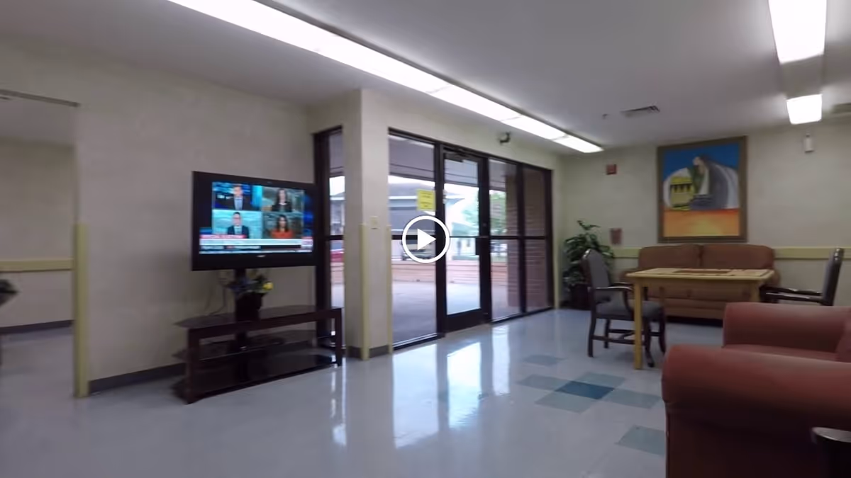 A nursing-home common area with a wall-mounted TV, seating including sofas and chairs around a table, and glass entry doors.