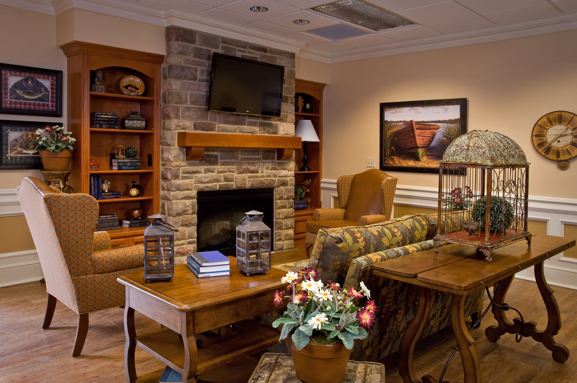 A cozy living room with a stone fireplace and a mounted flat-screen TV above it. The room features wooden bookshelves on either side of the fireplace filled with books and decorative items. There are two upholstered armchairs and a floral-patterned sofa arranged around a wooden coffee table with two lanterns and a stack of books. A wooden console table behind the sofa holds a decorative birdcage and a potted plant with flowers. The walls are adorned with framed artwork and a round clock.
