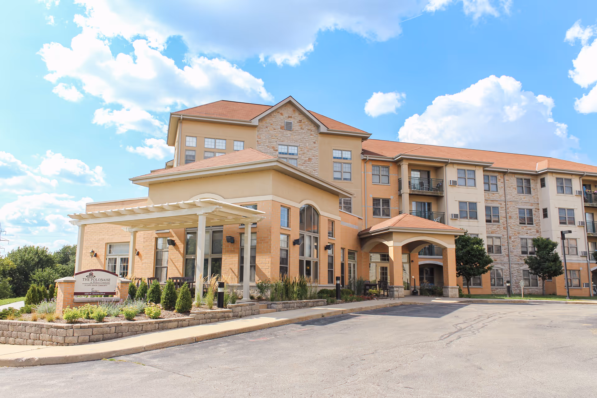 Exterior view of a multi-story assisted living facility building with a covered entrance and landscaped greenery under a partly cloudy blue sky.