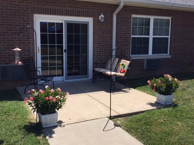 Small concrete patio outside a brick retirement community unit with a sliding glass door, two chairs, potted flowering plants, and a bird feeder.